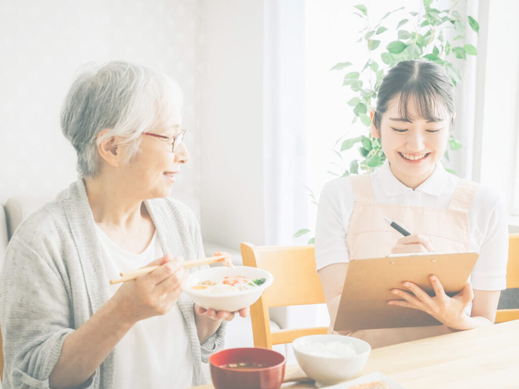 看護師が利用者の女性と笑顔で会話しながら食事の記録を取る様子